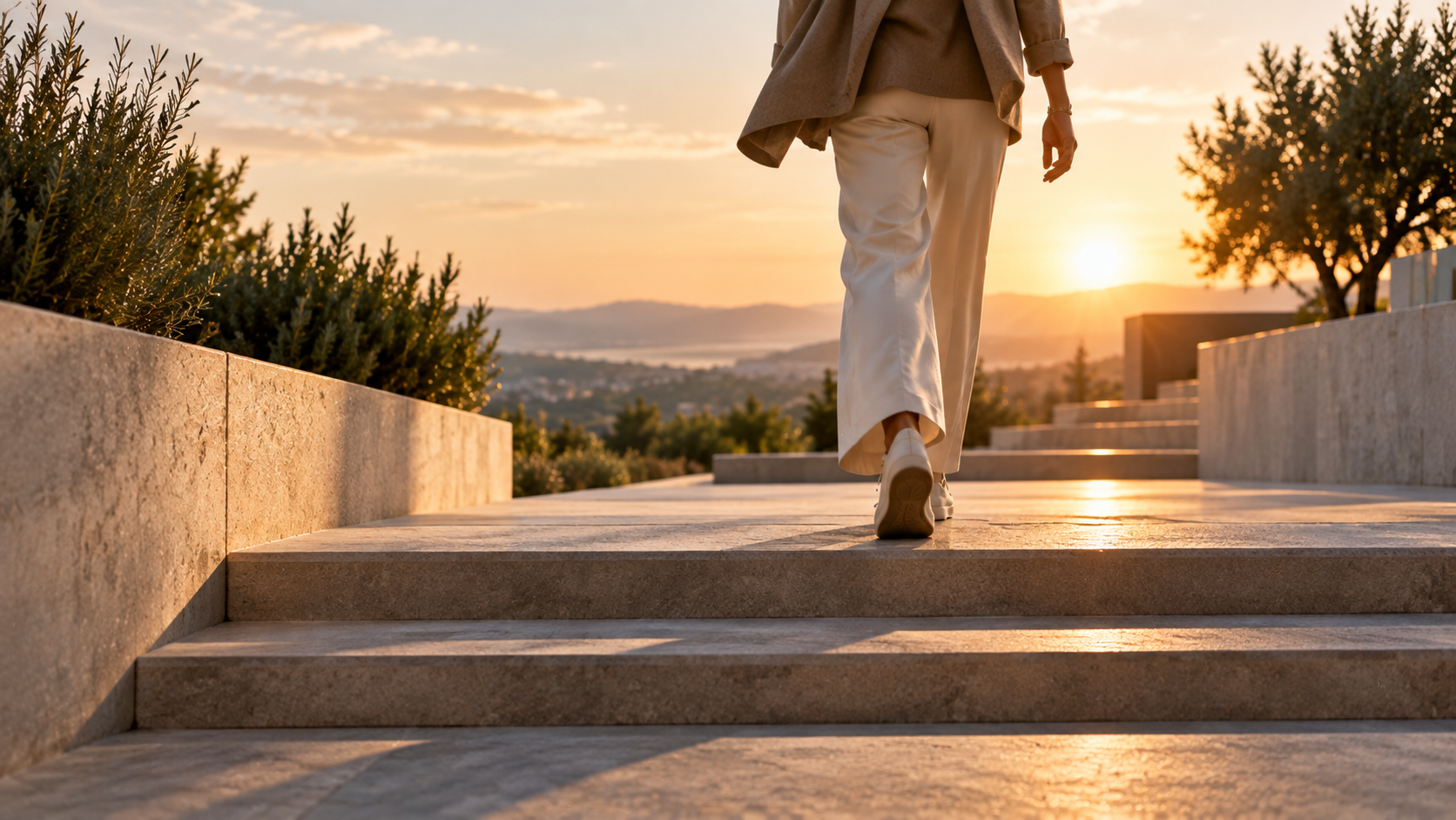 Woman in a beige coat walking up stone steps toward the sunset